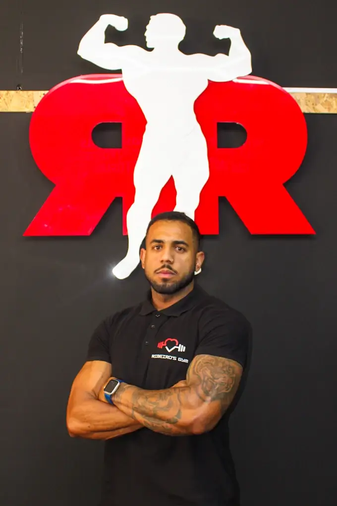 Ruan Ribeiro de braços cruzados, com o polo do Ribeiro's Gym, em frente ao monograma RR na parede do estúdio