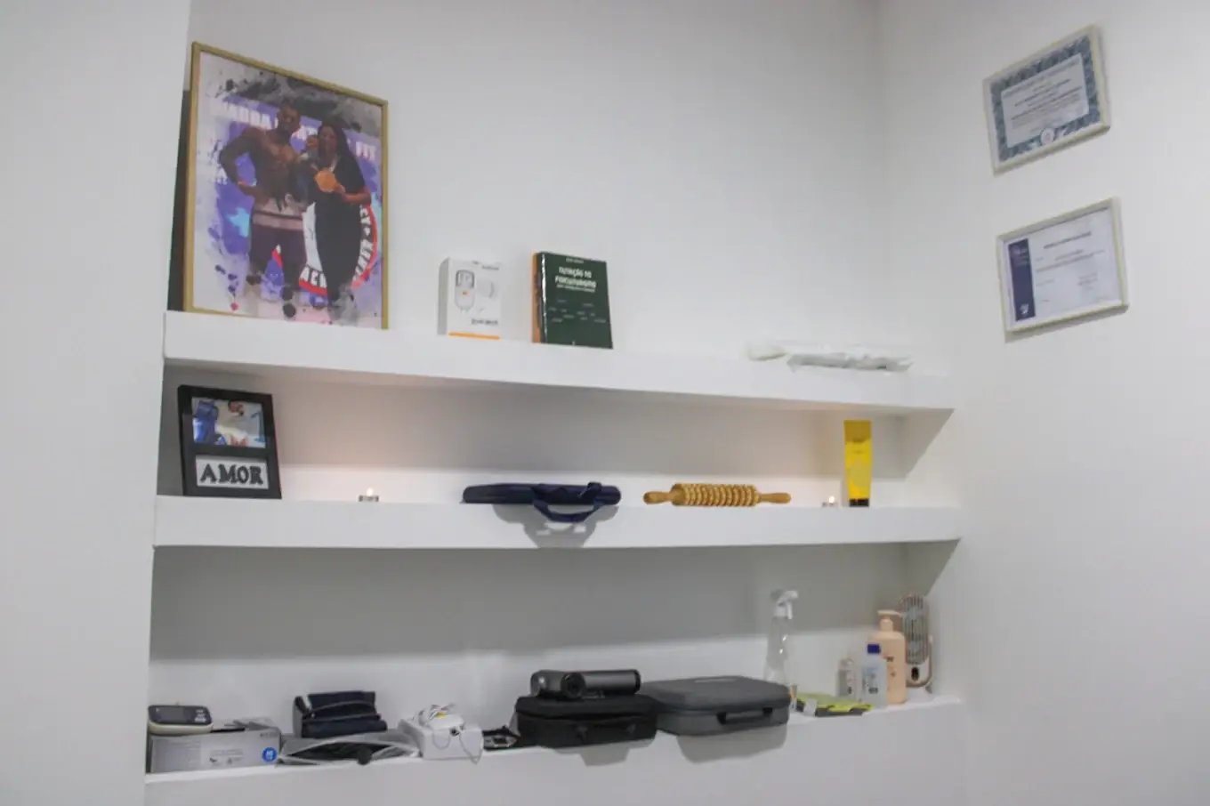 Sala de osteopatia branca com prateleiras, material de terapia manual e certificados emoldurados